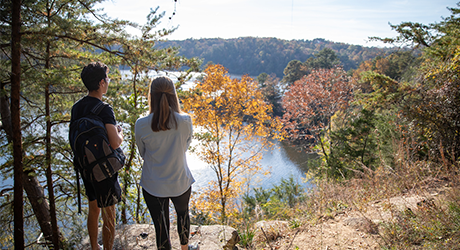 A man and a woman standing on a trail looking out to the distance of the forest. The sun is out and gleaming off the lake below.